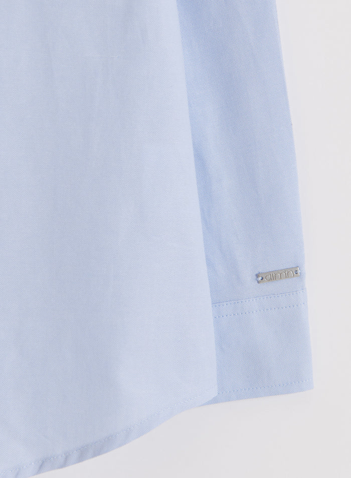 Flat lay close-up of a blue aim'n Oxford Shirt, showing the sleeve and a metal aim'n logo tag on the cuff.