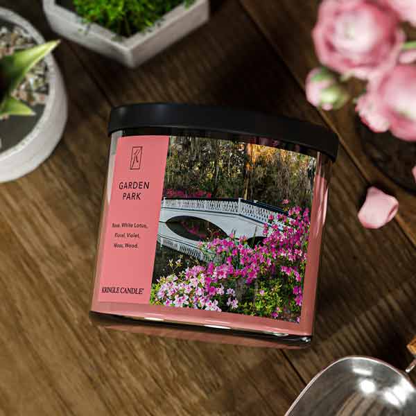 A three-wick candle in a clear glass jar with a black lid and a serene garden-themed label. The label features a white bridge arching over a pond, surrounded by blooming pink flowers and lush greenery. The candleβs wax is a soft pink shade, and the wicks are unlit. The scent notes listed are rose, white lotus, floral, violet, moss, and wood.