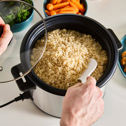 Person using a rice cooker with cooked rice inside, surrounded by vegetables on a table.