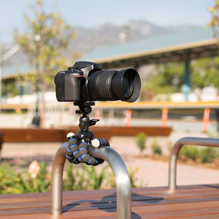 The 11 Inch Flexible Camera Tripod for Canon, Nikon, Samsung, and other 1/4-20 digital cameras is wrapped around an outdoor metal rail, holding a DSLR camera with benches, greenery, and blurred buildings in the background.