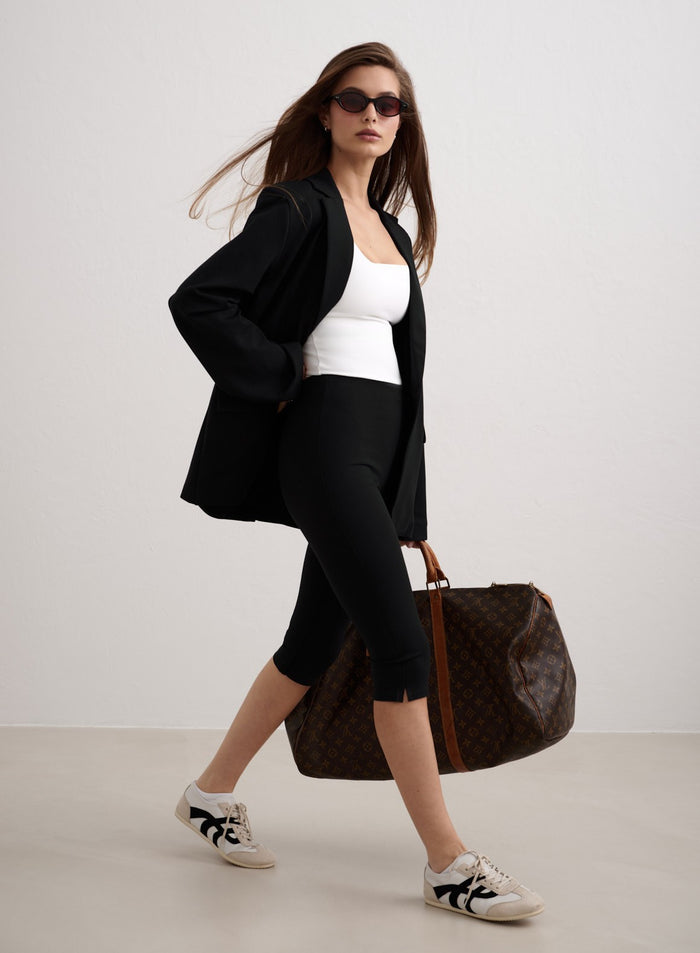Woman in black Luxe Capri Pants and a blazer walking with a large travel bag. [34-174cm]
