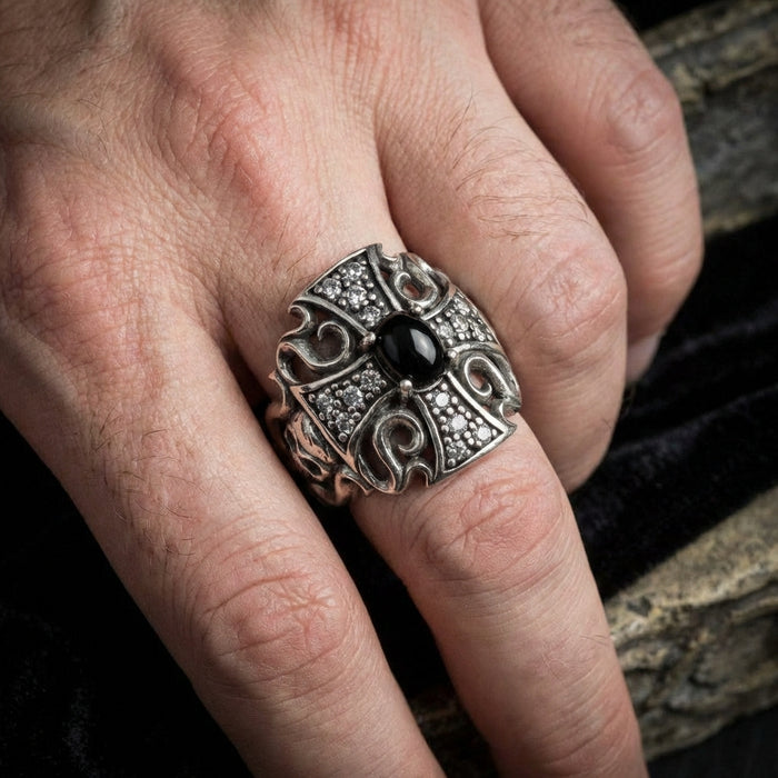Close-up of a man's hand wearing an ornate silver gothic cross ring featuring a central black oval stone surrounded by small clear crystals, set against a dark textured background.