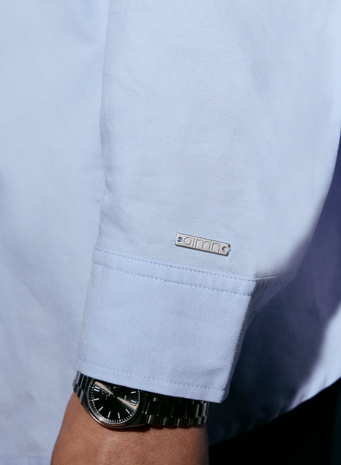 Close-up of a silver aimn logo tag on the cuff of a blue oxford shirt, worn by a person with a watch [M-170cm]