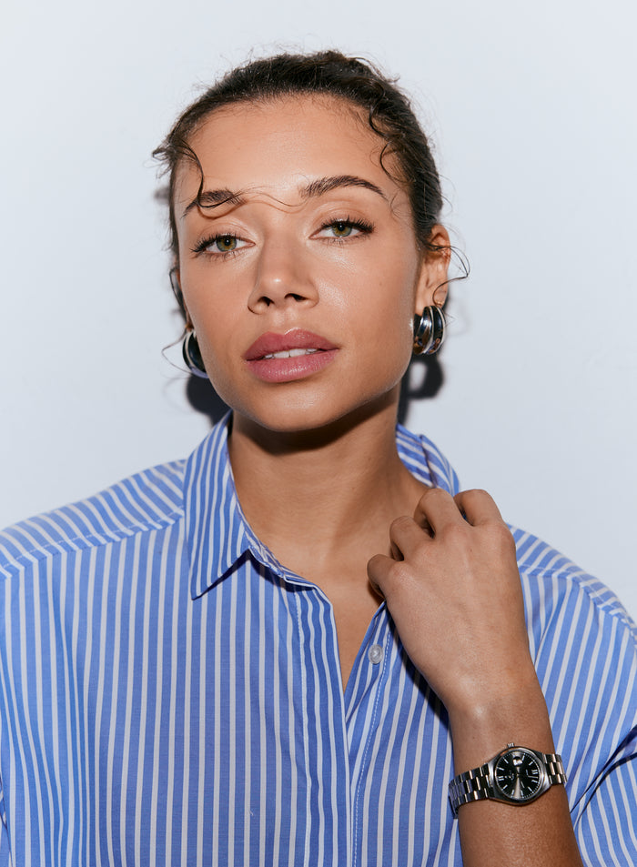 A woman wearing a blue stripe poplin shirt, looking forward, with her hand near her collar [M-170cm]