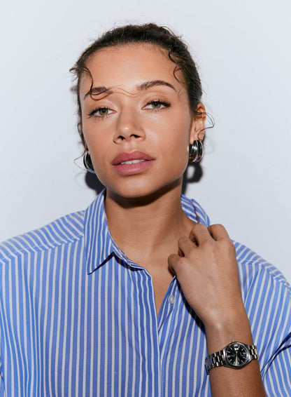 A woman wearing a blue stripe poplin shirt, looking forward, with her hand near her collar [M-170cm]