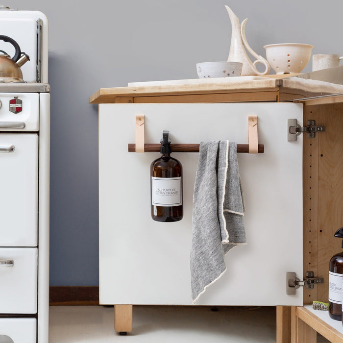 Cabinet Rail installed on a white kitchen cabinet door with vegetable-tan leather straps and a birch dowel holding a towel and cleaning bottle, showcasing Keyaiira’s space-saving under-sink storage solution.