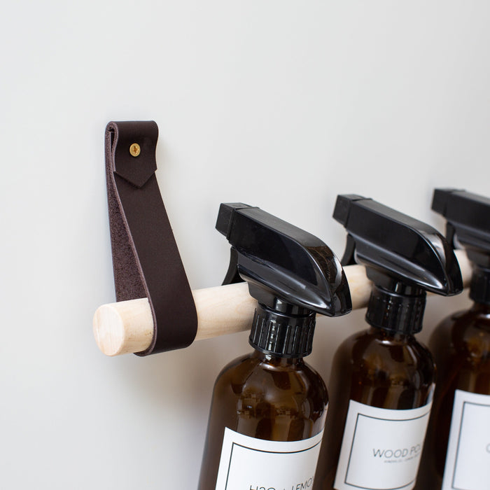 Chocolate leather Cabinet Rail with birch dowel mounted on the inside of a cabinet door, storing amber spray bottles for organized under-sink cleaning supply storage.