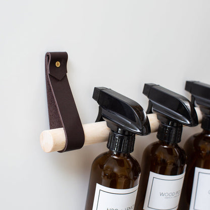 Chocolate leather Cabinet Rail with birch dowel mounted on the inside of a cabinet door, storing amber spray bottles for organized under-sink cleaning supply storage.