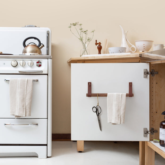 Keyaiira brown leather dowel rack shown in a neutral kitchen interior, used to hang towels and utensils. Modern handcrafted wall-mounted kitchen storage solution.