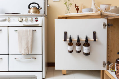 Keyaiira handcrafted leather dowel rack in black used inside a kitchen cabinet to hold refillable amber spray bottles. Functional leather strap storage solution with natural wood dowel, perfect for organizing cleaning products in modern minimalist kitchens.