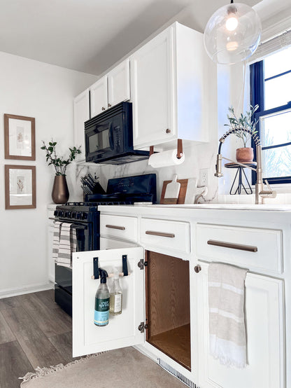 Bright modern kitchen featuring Keyaiira leather dowel racks in multiple finishes used for paper towel and bottle storage. Handcrafted leather and wood hanging rails add practical style to white cabinetry and minimalist home decor.