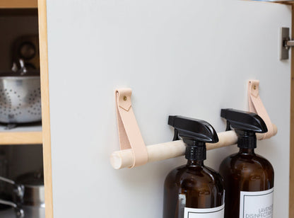 Under-sink storage solution using Keyaiira handcrafted vegetable-tanned leather straps and wooden dowel to organize cleaning bottles. Simple, modern, and durable home organization accessory.