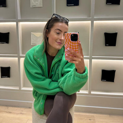Woman taking mirror selfie holding iPhone with pink orange checkered iPhone case, wearing green fleece in store