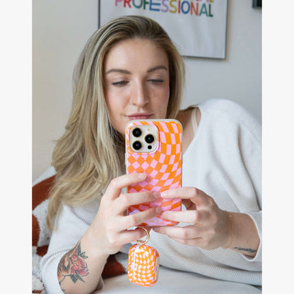 Woman taking a selfie while holding an iPhone with a pink and orange checkered case and matching AirPods case on a keyring
