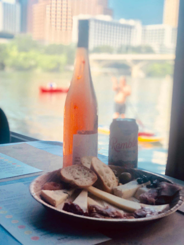 Image of cheese plate, bottle of wine, rambler on a boat with kayaker in the background