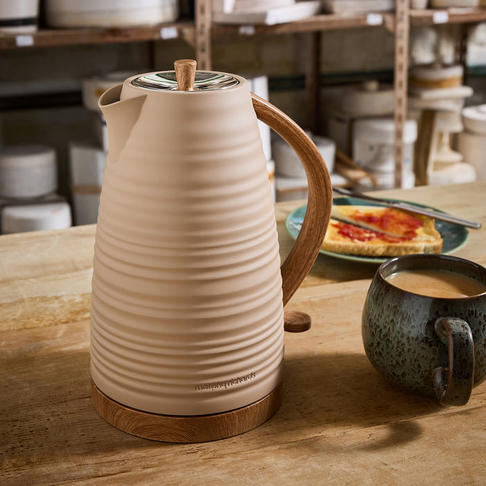 Beige ceramic kettle with wooden handle on a wooden table, with a cup of coffee and a plate of toast in the background.