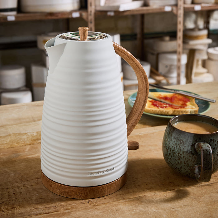 White ceramic kettle with wooden handle on a wooden table, with a cup of coffee and plate of food in the background.