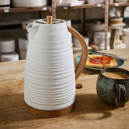 White ceramic kettle with wooden handle on a wooden table, with a cup of coffee and plate of food in the background.