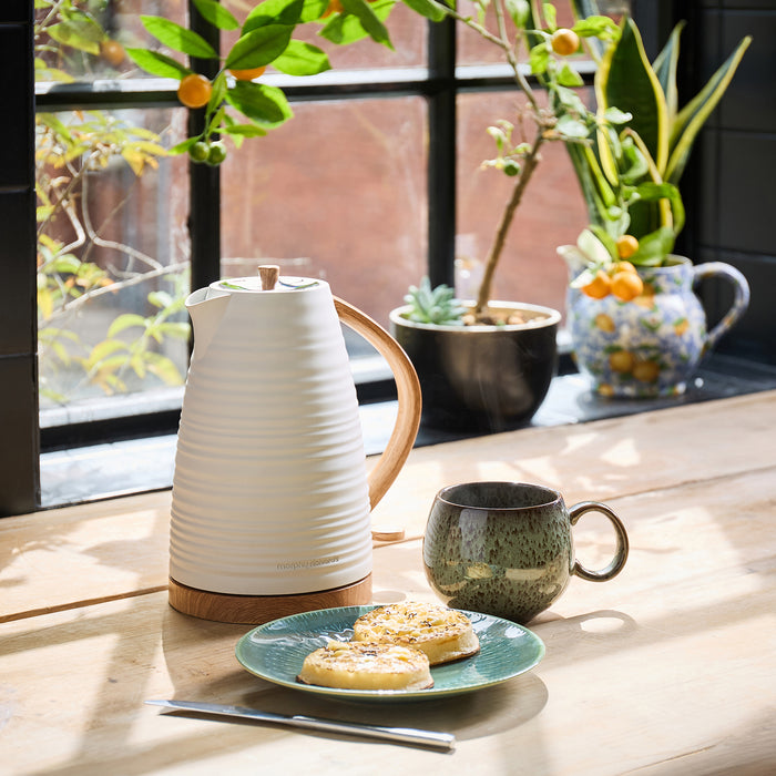 White ceramic Kettle, green mug, and plate with cookies on a wooden table with plants in the background.