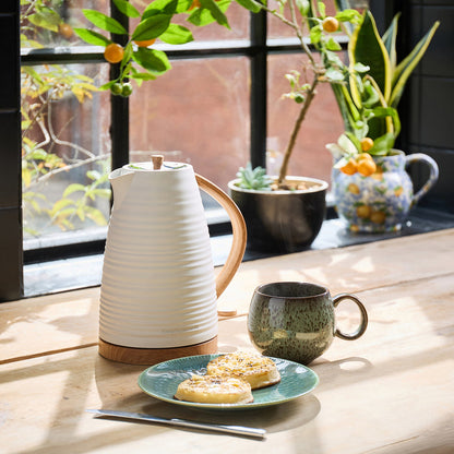 White ceramic Kettle, green mug, and plate with cookies on a wooden table with plants in the background.