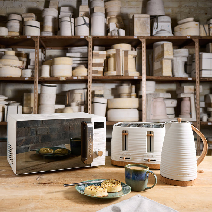 Kitchen appliances including a microwave, toaster, and kettle on a wooden table with shelves of kitchenware in the background.