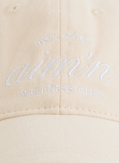 Sand cotton Club Cap with a white embroidered aim'n logo resting on a silver metal chair.