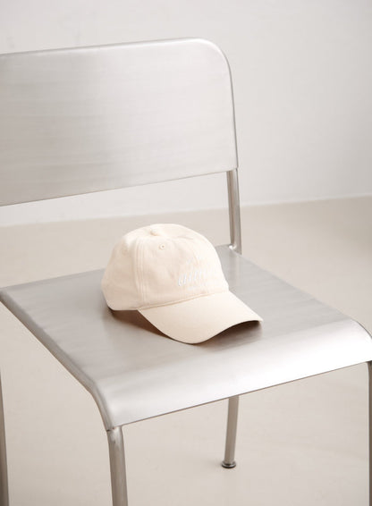 Sand-colored cotton Club Cap with white embroidered aim'n logo resting on a minimalist silver chair.