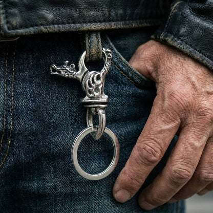 Close-up of a rugged silver flame dragon keychain clipped to the belt loop of blue denim jeans, worn by a man in a black leather jacket with his hand resting near the pocket.