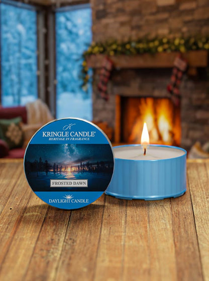 Kringle Candle Frosted Dawn daylight candle with packaging on a wooden surface in front of a fireplace.