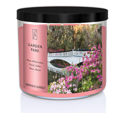 A three-wick candle in a clear glass jar with a black lid and a serene garden-themed label. The label features a white bridge arching over a pond, surrounded by blooming pink flowers and lush greenery. The candle’s wax is a soft pink shade, and the wicks are unlit. The scent notes listed are rose, white lotus, floral, violet, moss, and wood.