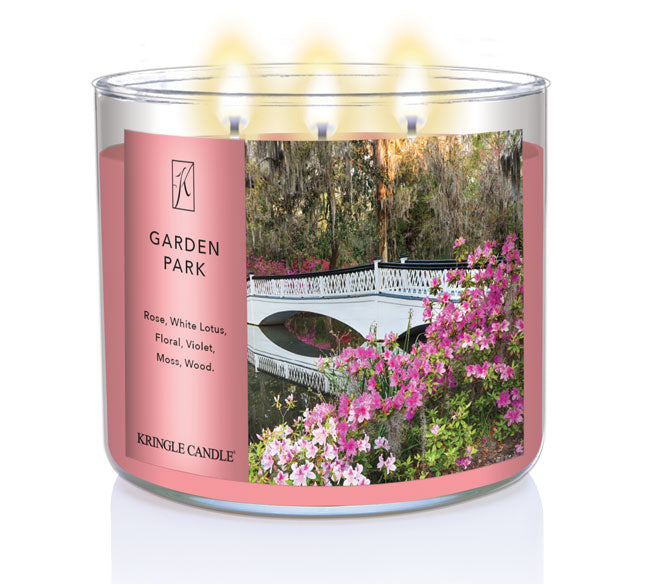 A three-wick lit candle in a clear glass jar with a serene garden-themed label. The candle’s wax is a soft pink shade, and all three wicks are burning.