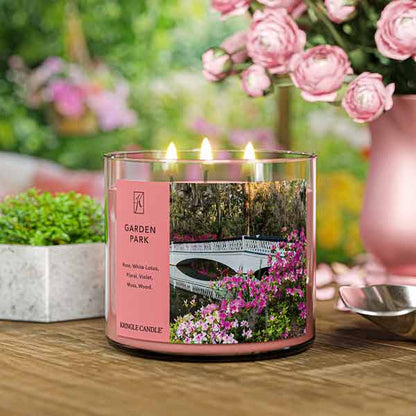 A three-wick lit candle in a clear glass jar with a serene garden-themed label. The candle’s wax is a soft pink shade, and all three wicks are burning.