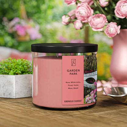 A three-wick candle in a clear glass jar with a black lid and a serene garden-themed label. The label features a white bridge arching over a pond, surrounded by blooming pink flowers and lush greenery. The candle’s wax is a soft pink shade, and the wicks are unlit. The scent notes listed are rose, white lotus, floral, violet, moss, and wood.