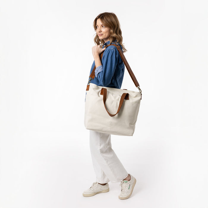 Off-white canvas tote bag displayed with navy blue and warm brown leather accents, styled with cream and neutral tones