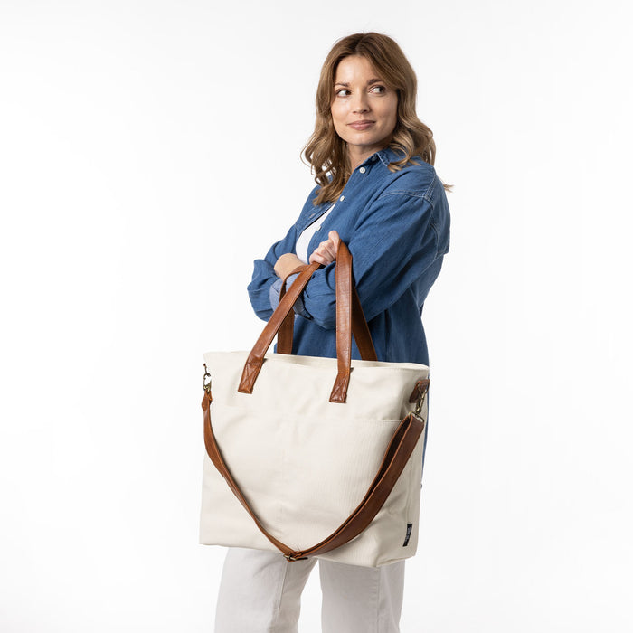 Cream canvas tote bag with cognac brown leather handles and trim, displayed on a model wearing a white long-sleeve top