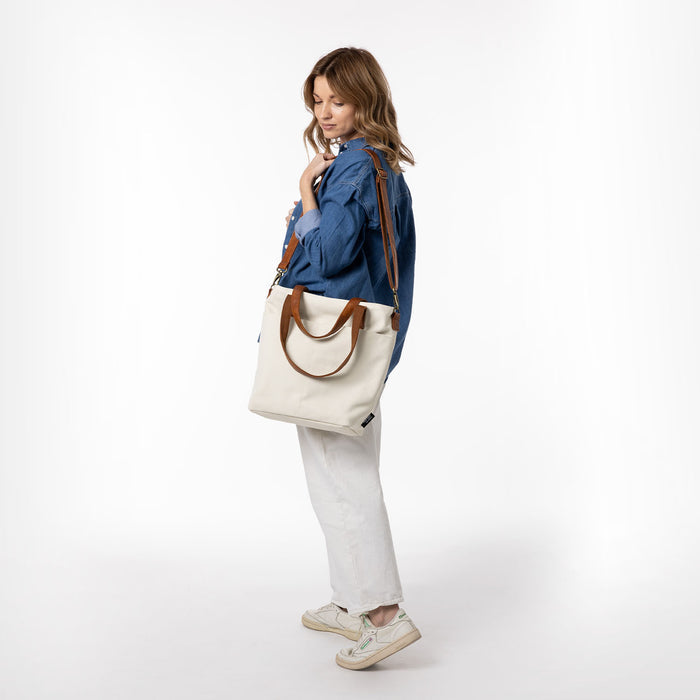 Cream canvas tote bag held by person wearing white pants and neutral sneakers against a neutral background