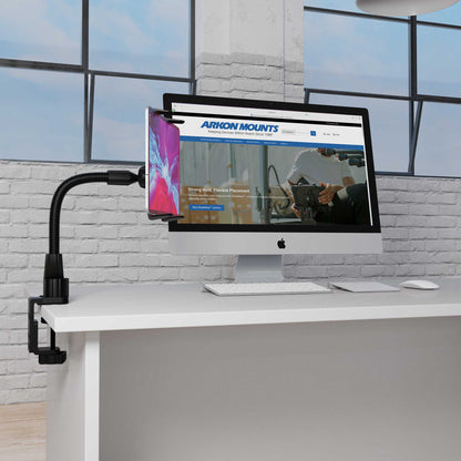 The Heavy-Duty Slim-Grip® Tablet Clamp Mount secures a smartphone to a white desk next to an iMac showing the Arkon Mounts website in a modern office with large windows and a white brick wall.