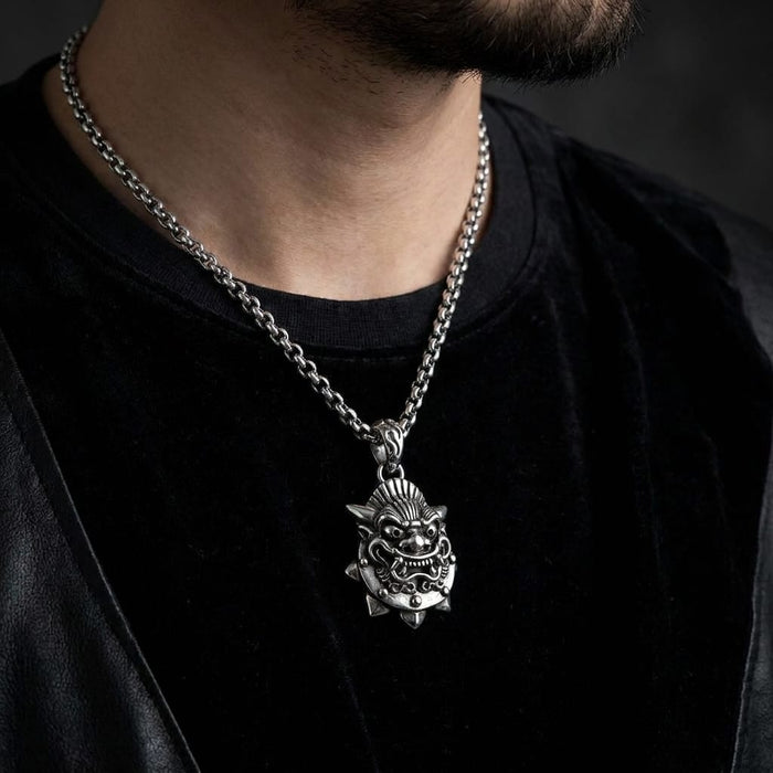 Close-up of a bearded male model wearing a detailed sterling silver Japanese demon mask pendant on a thick silver chain, styled with a black t-shirt and leather jacket against a dark, moody background.