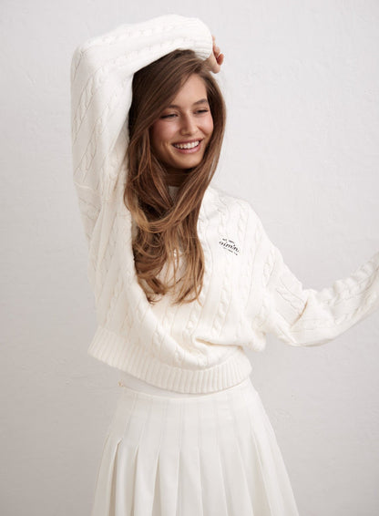 Off-white cotton cable-knit sweater by aim'n styled with a white pleated skirt on a smiling model. [M-174cm]