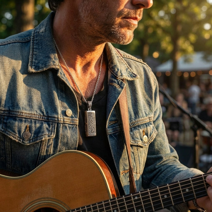 Silver mini harmonica pendant necklace with ornate engraved design worn by a man in a denim jacket playing acoustic guitar at an outdoor concert setting