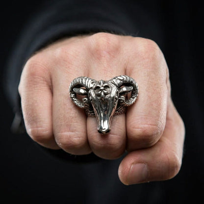 Close-up of a man's clenched fist wearing a bold sterling silver ram head biker ring featuring curved horns and a fleur-de-lis detail on the forehead, set against a dark background.