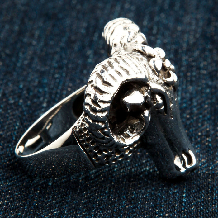 Detailed view of the heavy silver ram head ring resting on dark denim, emphasizing the curled ridged horns and textured band.