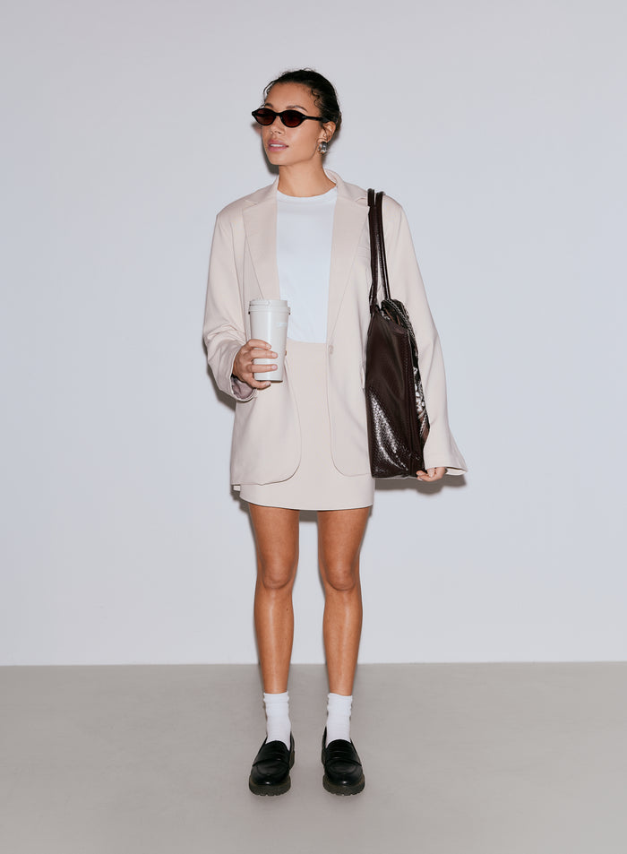 A woman wearing a sand colored Luxe Relaxed Blazer, a white t-shirt, and a matching skirt, holding a reusable coffee cup and a dark brown tote bag. [40-170CM]