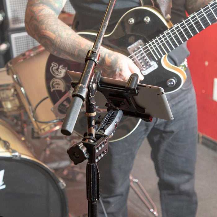 A tattooed musician plays an electric guitar by a drum set. In front of them, a Slim-Grip® Tablet Holder with Heavy-Duty Clamp Mount secures a tablet on a stand, likely displaying music or lyrics in the rehearsal space.