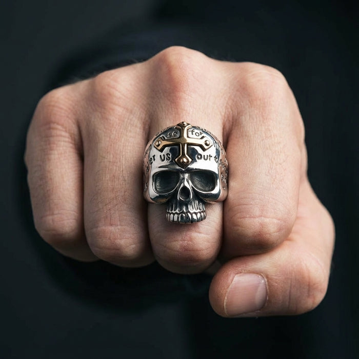 Close-up of a heavy sterling silver skull ring featuring a gold cross accent on the forehead and engraved lettering, worn on a man's clenched fist against a dark background.