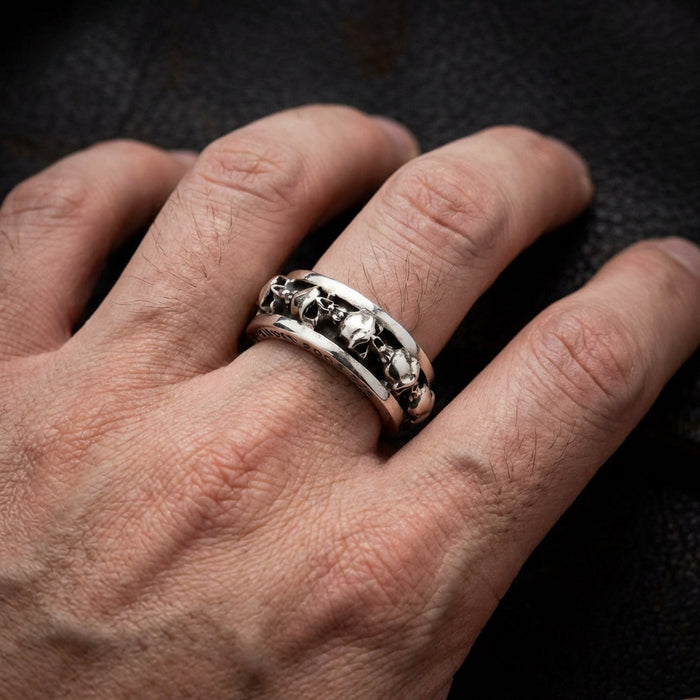 Sterling silver gothic skull ring with multiple skull designs worn on a man's finger against a dark textured background, showcasing the detailed craftsmanship of this edgy biker-style stacking band