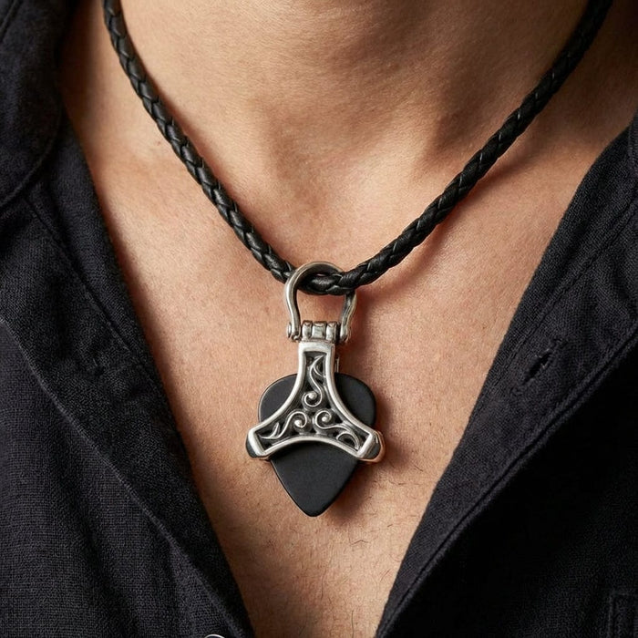 Silver tribal guitar pick holder pendant with ornate Celtic-style scrollwork design holding a black guitar pick, worn on a braided black leather cord necklace against a man's chest with open black shirt