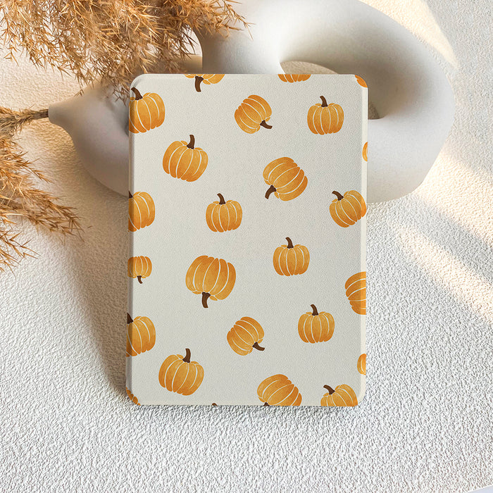 A Kindle case with a watercolor pumpkin pattern on a beige background, displayed against a backdrop of dried plants and a white abstract sculpture.