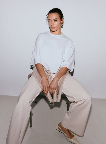 A woman wearing a white Luxe Boxy Short Sleeve top, light beige wide-leg pants, and light beige flats, sitting on a chair with a sweater draped over her lap. [M-170cm]