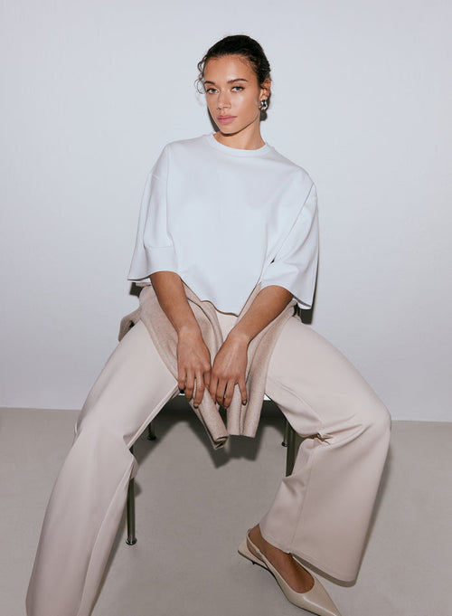 A woman wearing a white Luxe Boxy Short Sleeve top, light beige wide-leg pants, and light beige flats, sitting on a chair with a sweater draped over her lap. [M-170cm]